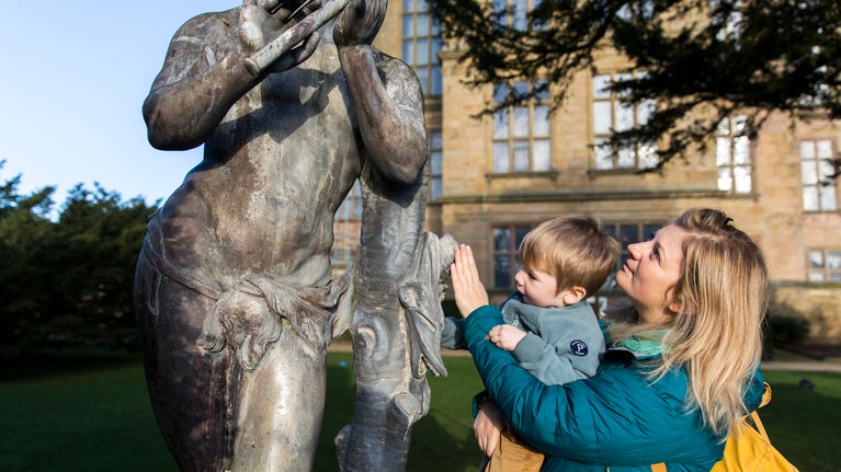 A mother with a teal coat and bright yellow backpack, holding her young son with light brown hair, reaches for a grey statue in the gardens, with the sand-coloured Hardwick Hall seen in the backdrop.